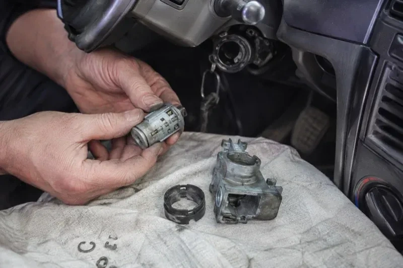 Hands holding a removed ignition cylinder beside a car steering column, with the housing and small parts on a cloth. — Ignition Lock Repair, Hamilton